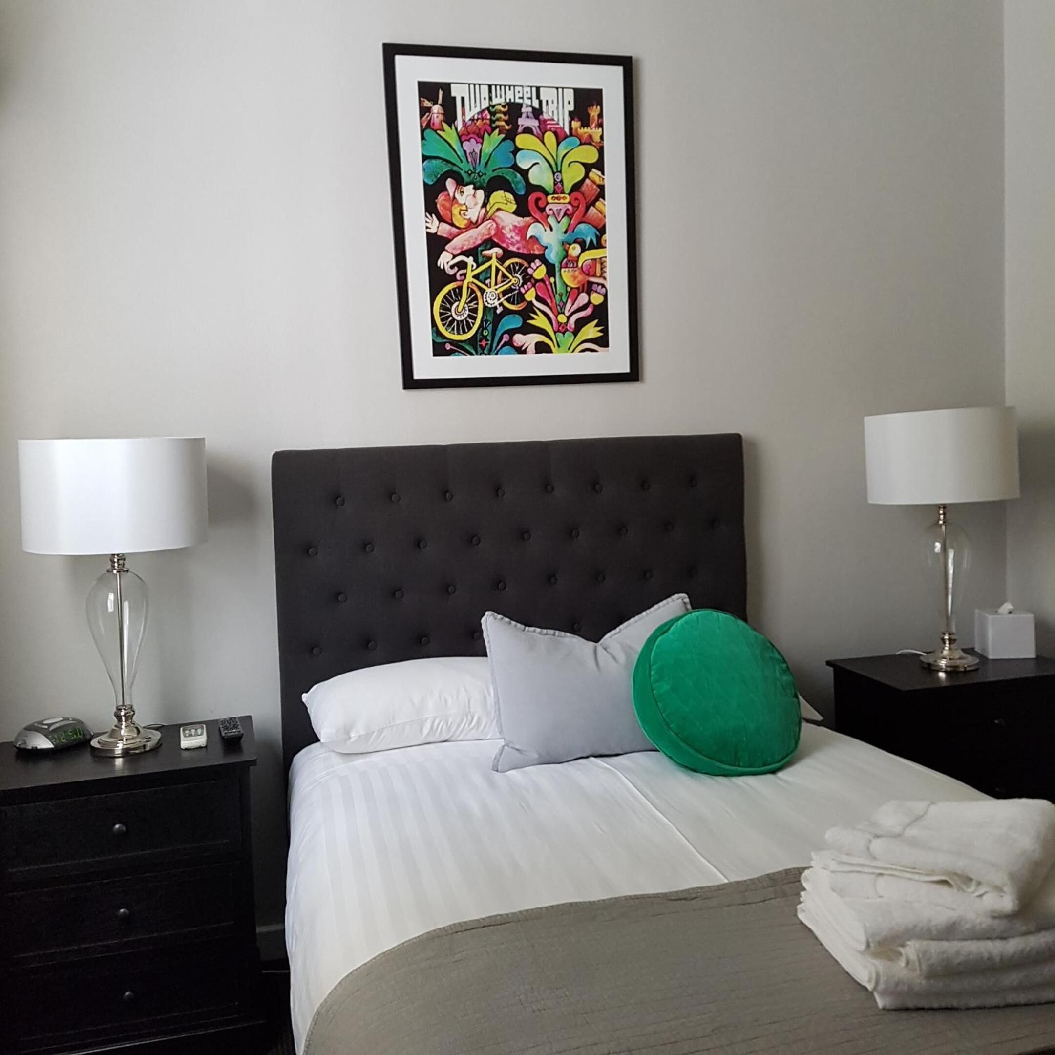 A neatly made bed with white linens, a grey headboard, and a colorful artwork above it. There are two bedside tables with lamps, a clock, and a remote control on the left side.