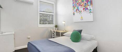 Ein Hotelzimmer mit Bett, Schreibtisch, Stuhl, Fenster mit Jalousien, einer Klimaanlage und einem farbenfrohen, abstrakten Gemälde an der Wand.
