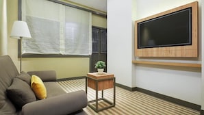 Una habitación de hotel moderna con un sofá, una pequeña mesita de noche con una planta, un televisor de pantalla plana grande montado en la pared y una ventana con cortinas translúcidas.