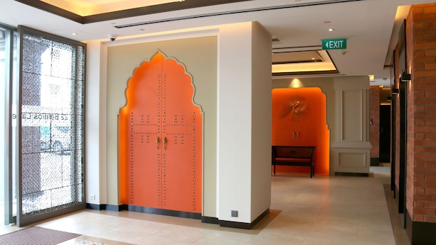 Un vestíbulo de hotel con una puerta grande de color naranja, un separador de vidrio con un diseño geométrico y un banco al fondo.