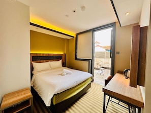 A hotel room with a bed, a desk, a chair, a small table, and a sliding door leading to a balcony.