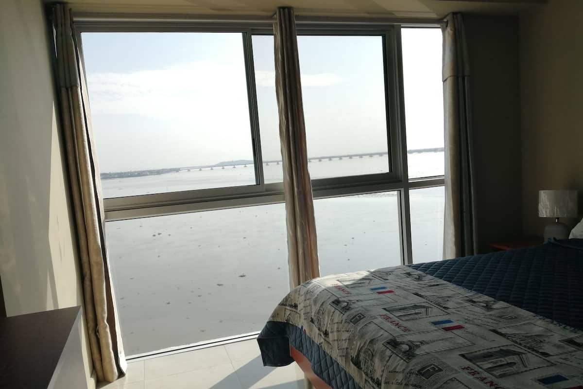 A hotel room with a large window overlooking a beach and a bridge. The room has a bed with a patterned bedspread and a nightstand with a lamp.