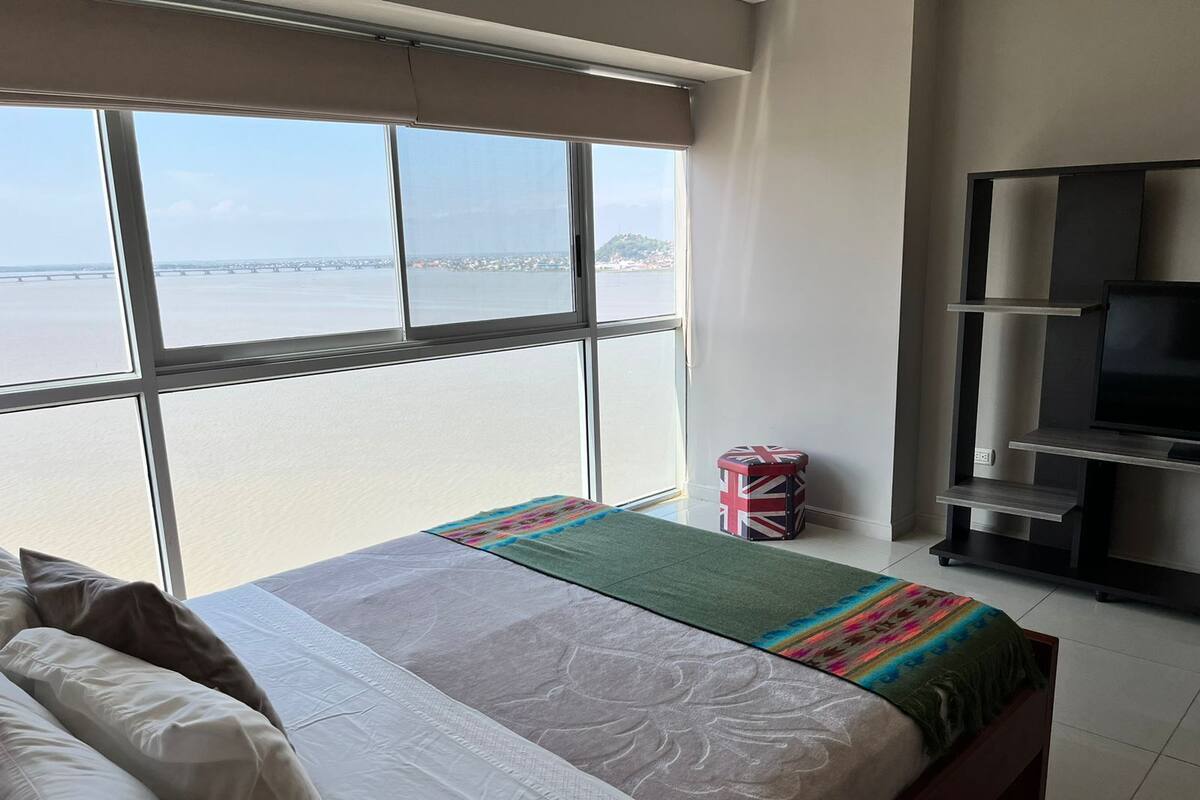 Ein Hotelzimmer mit einem großen Fenster, das einen Blick auf eine Brücke und Wasser freigibt. Es gibt ein Bett mit einer Decke in Grün und Rot, einen kleinen Hocker mit Union-Jack-Muster und eine TV-Konsole.