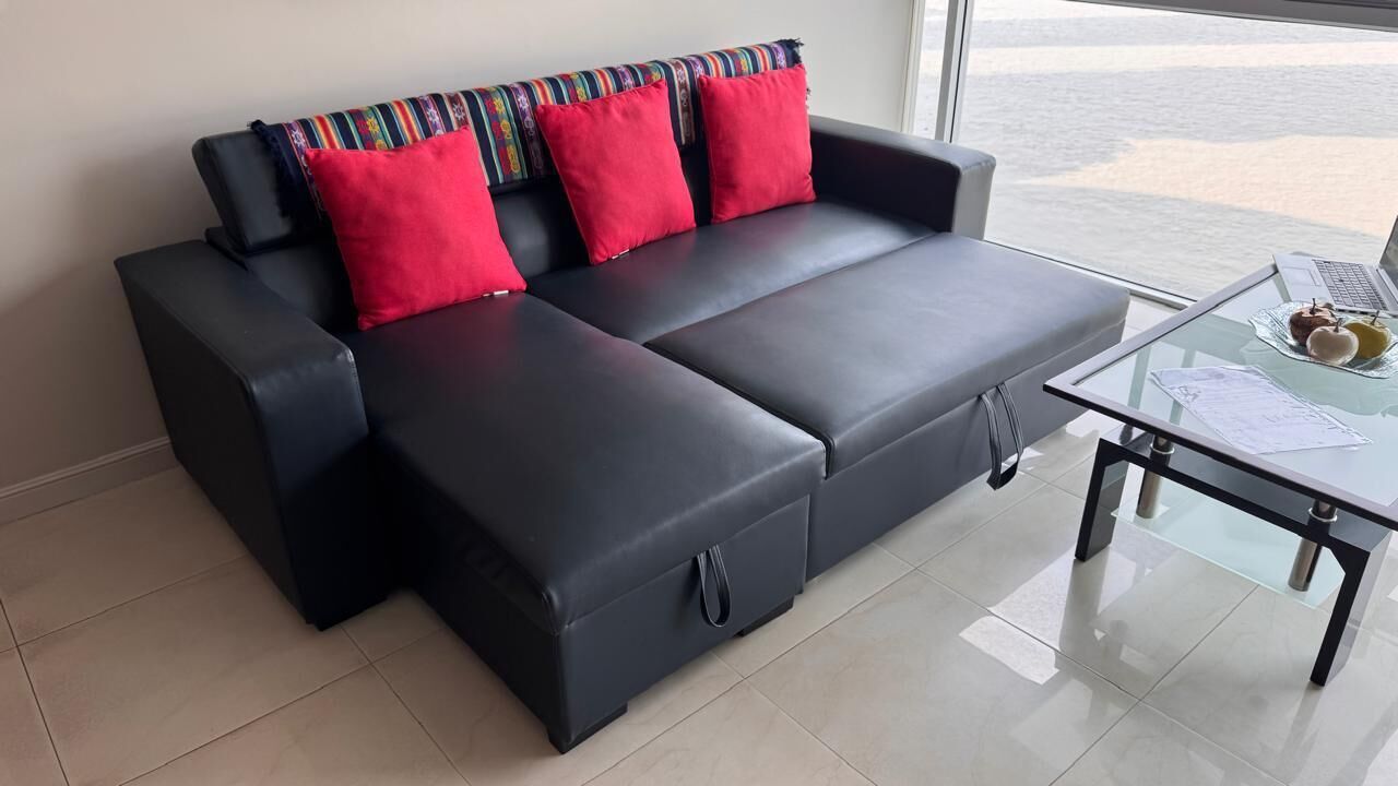 Un canapé d’angle noir avec des coussins rouges et une table basse en verre sur laquelle se trouvent un ordinateur portable et des en-cas.