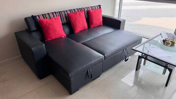Un canapé d’angle noir avec des coussins rouges et une table basse en verre sur laquelle se trouvent un ordinateur portable et des en-cas.