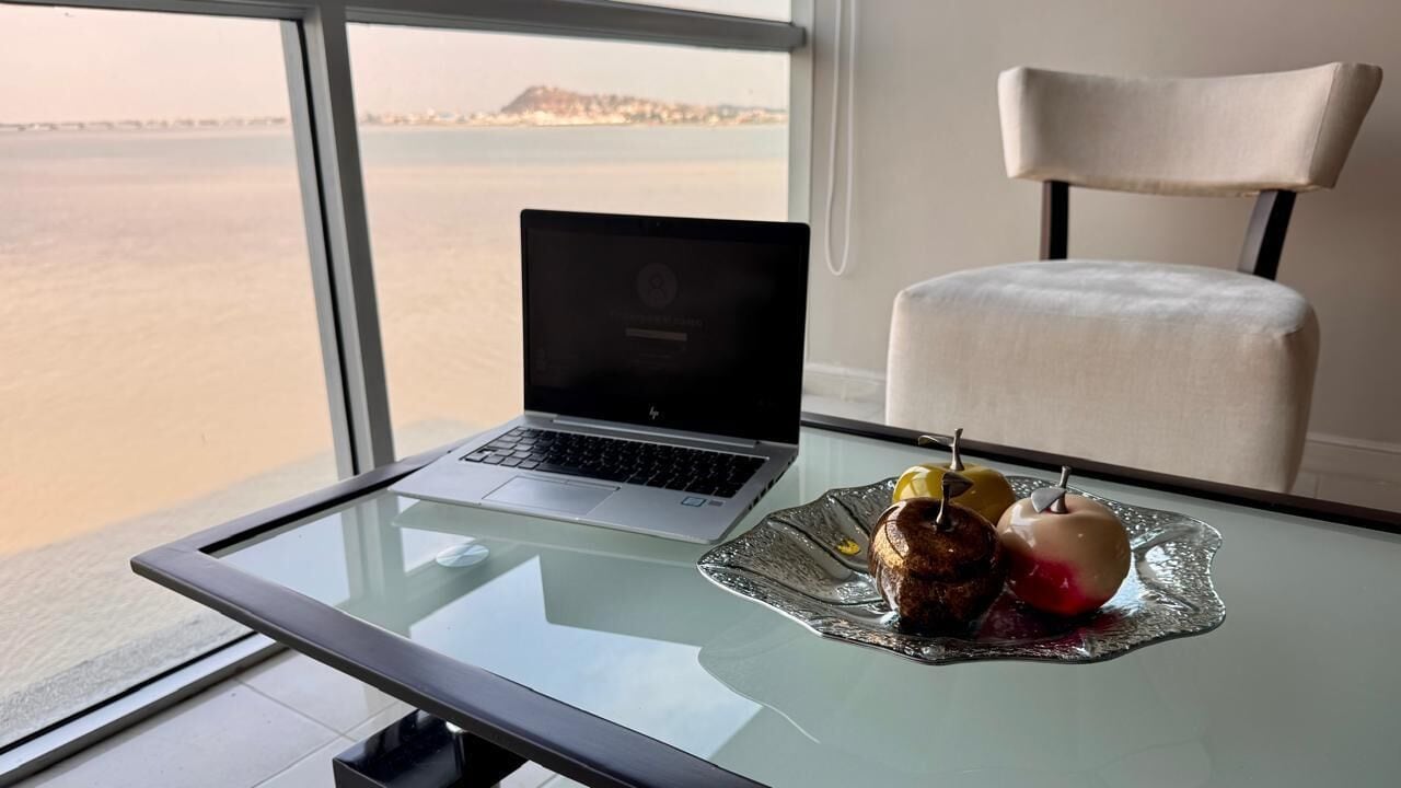 Un ordinateur portable est posé sur une table en verre, à côté d’une assiette de nourriture et d’une chaise, dans une pièce offrant une vue sur la mer.