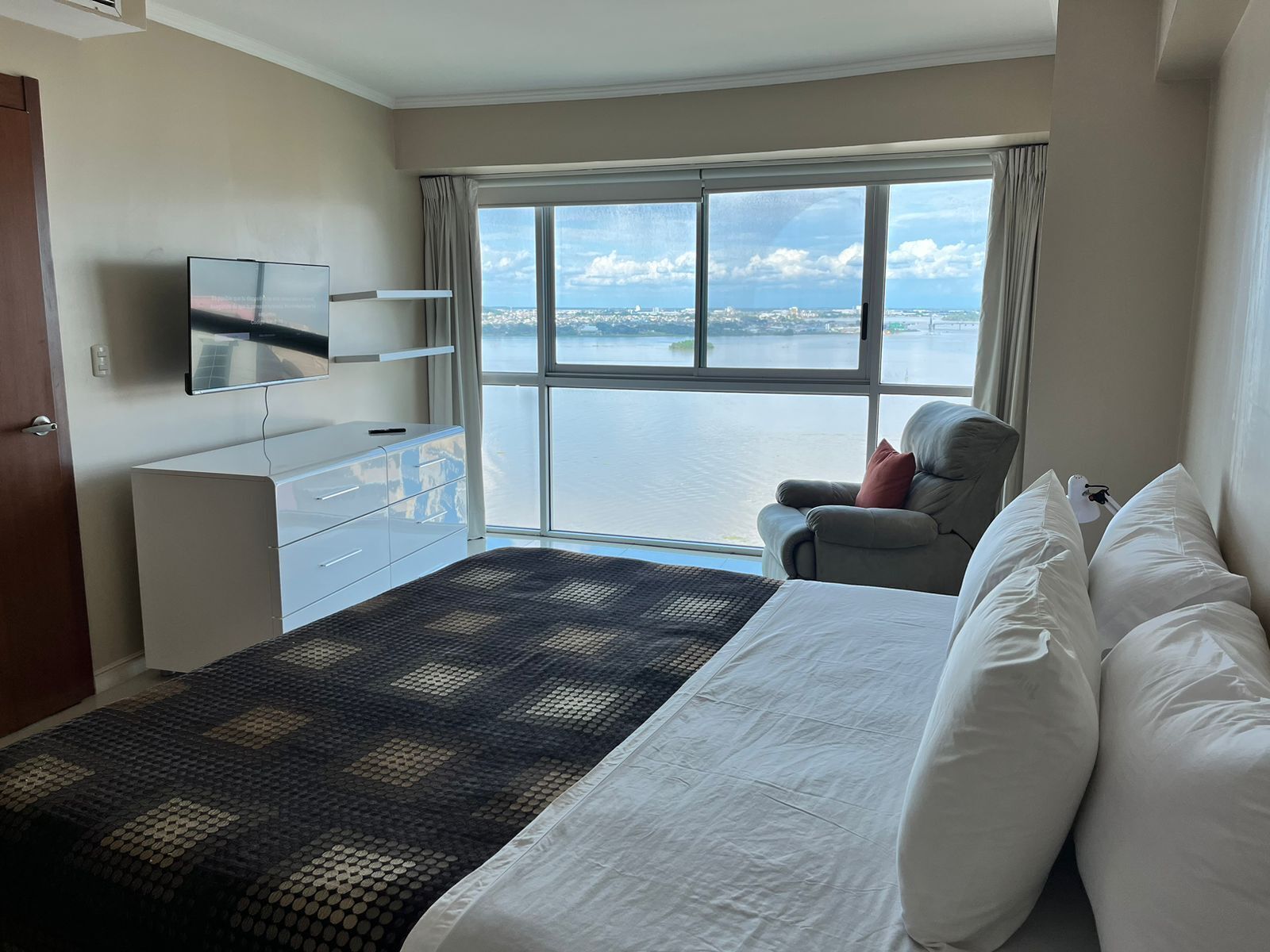 A hotel room with a bed, a chair, a TV, a dresser, and a large window offering a view of a body of water.