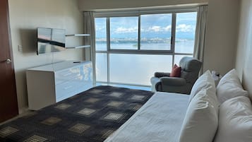 A hotel room with a bed, a chair, a TV, a dresser, and a large window offering a view of a body of water.