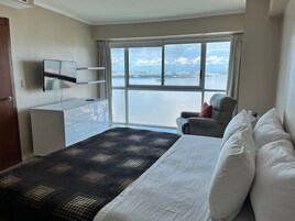 Habitación de hotel con cama, una silla, televisión, cómoda y un amplio ventanal con vista a una masa de agua.