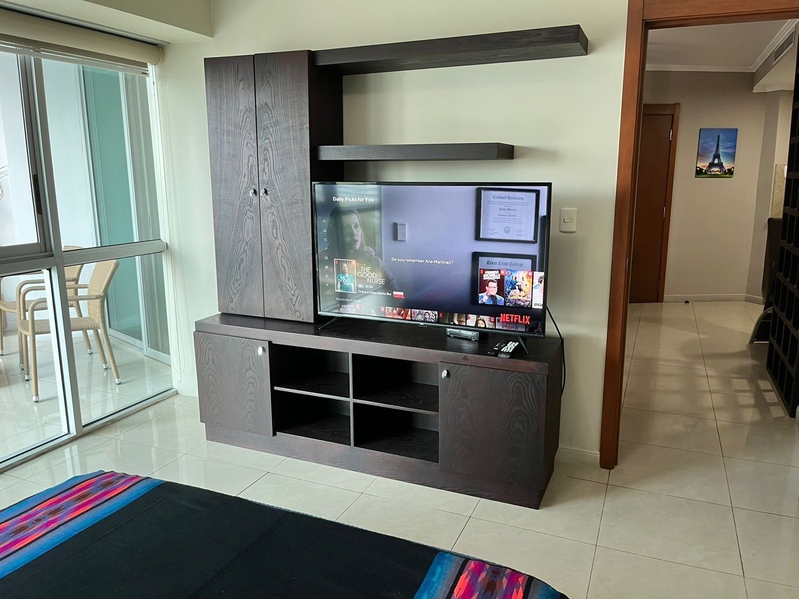 Sala de estar con televisor de pantalla plana montado en un mueble de madera oscura, una puerta de cristal que da a un balcón y una alfombra colorida en el suelo.