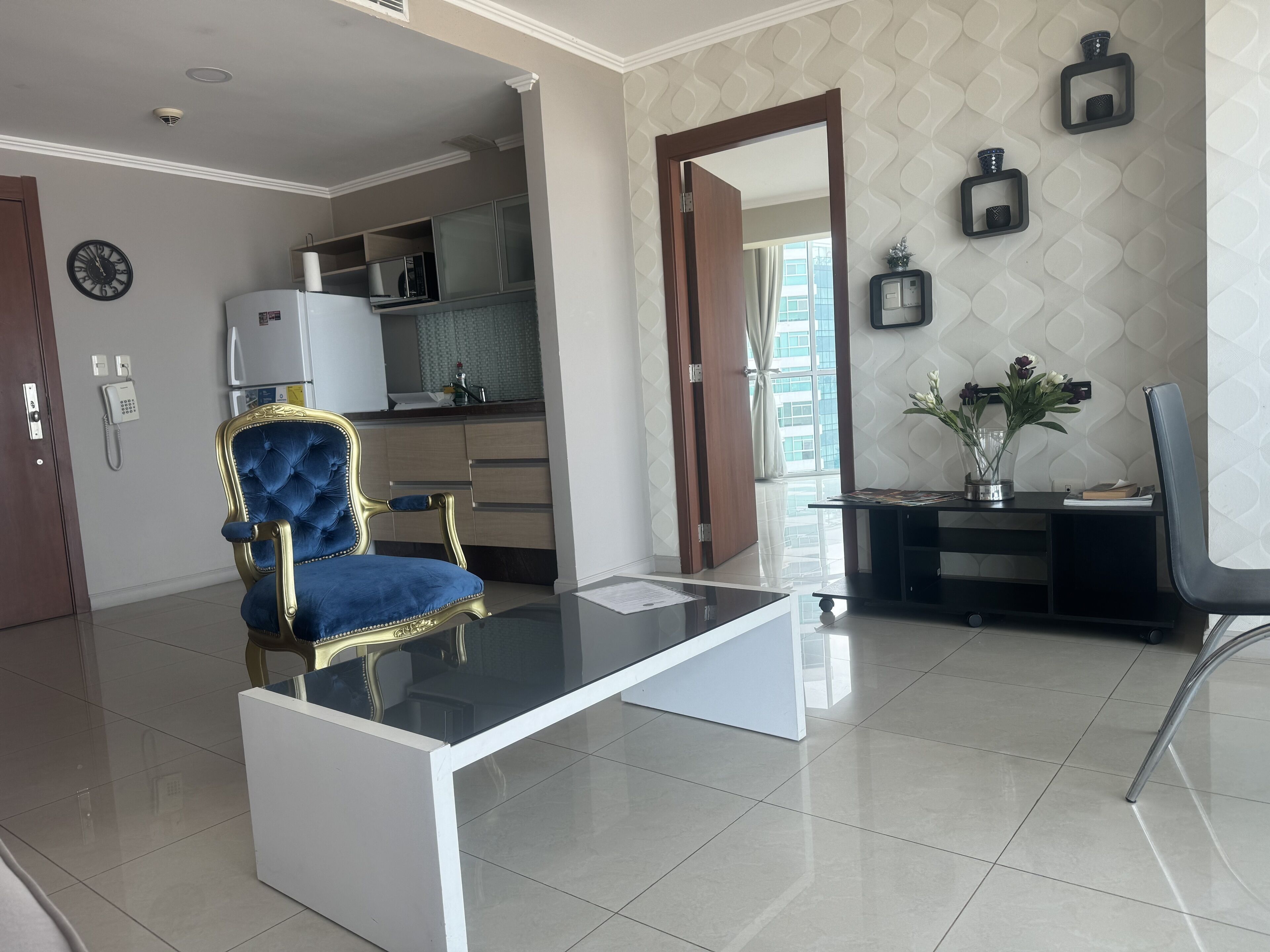 A modern living room with a glass coffee table, a blue upholstered armchair, a kitchen area with cabinets, and a decorative mirror.