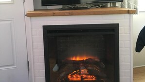 Ett modernt vardagsrum med en platt-TV på en trähylla ovanför en vit tegelkamin där man kan se elden.