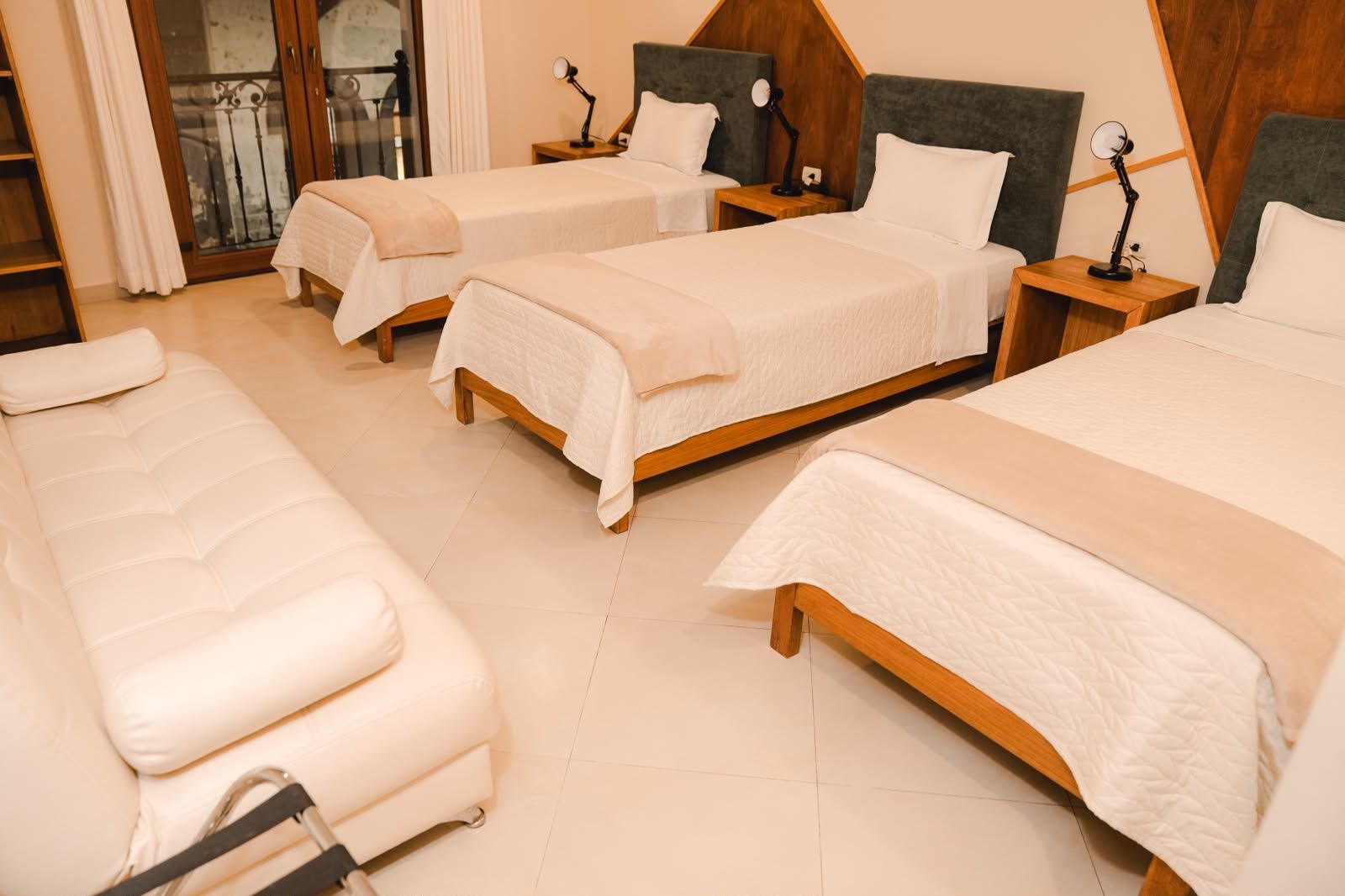 Habitación de hotel con tres camas, cabeceros de madera, mesitas de noche con lámparas y vista a un balcón.