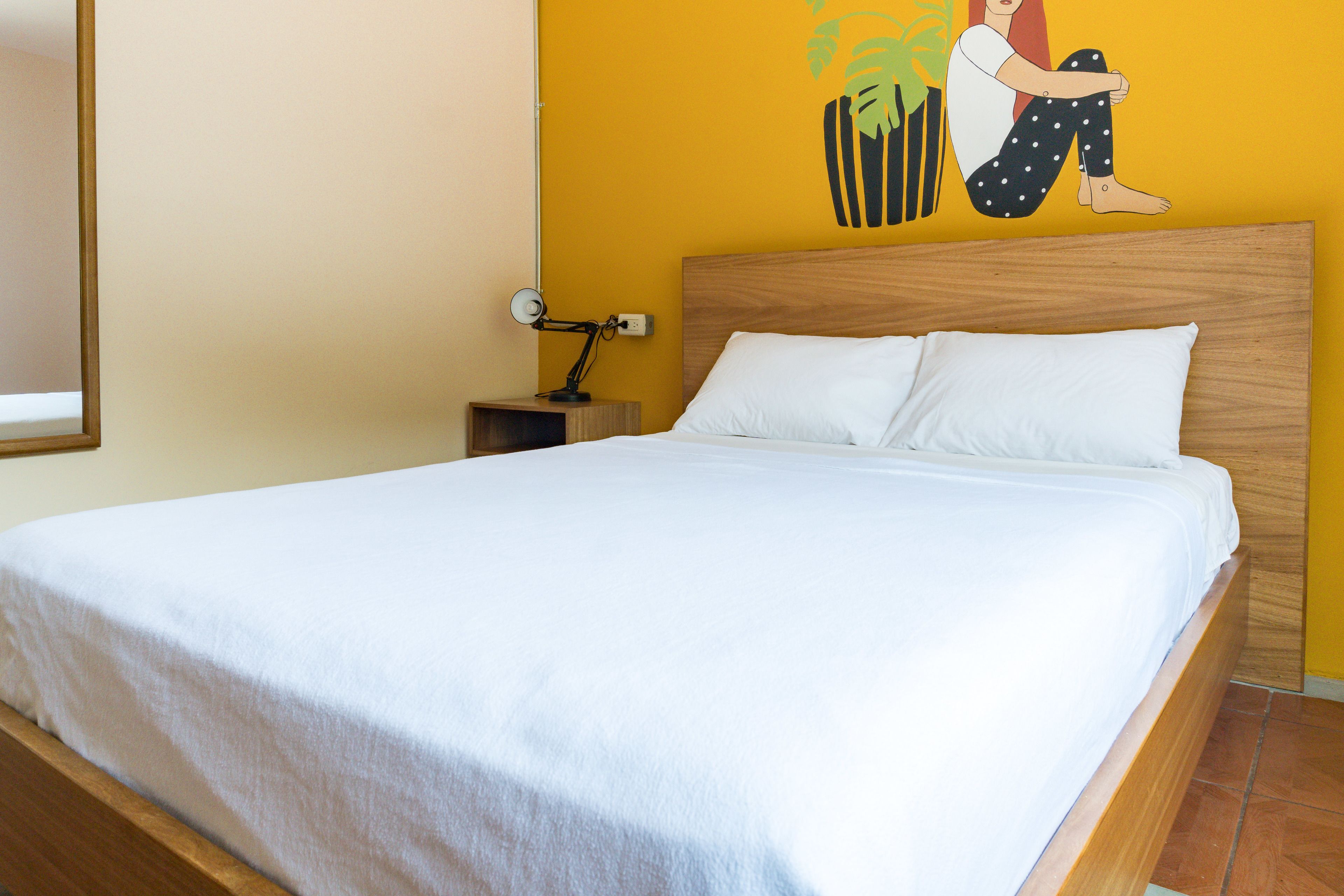 Un lit bien fait, avec du linge de lit blanc, dans une chambre dotée d’une tête de lit en bois, d’une petite table de chevet avec une lampe et d’une fresque murale.