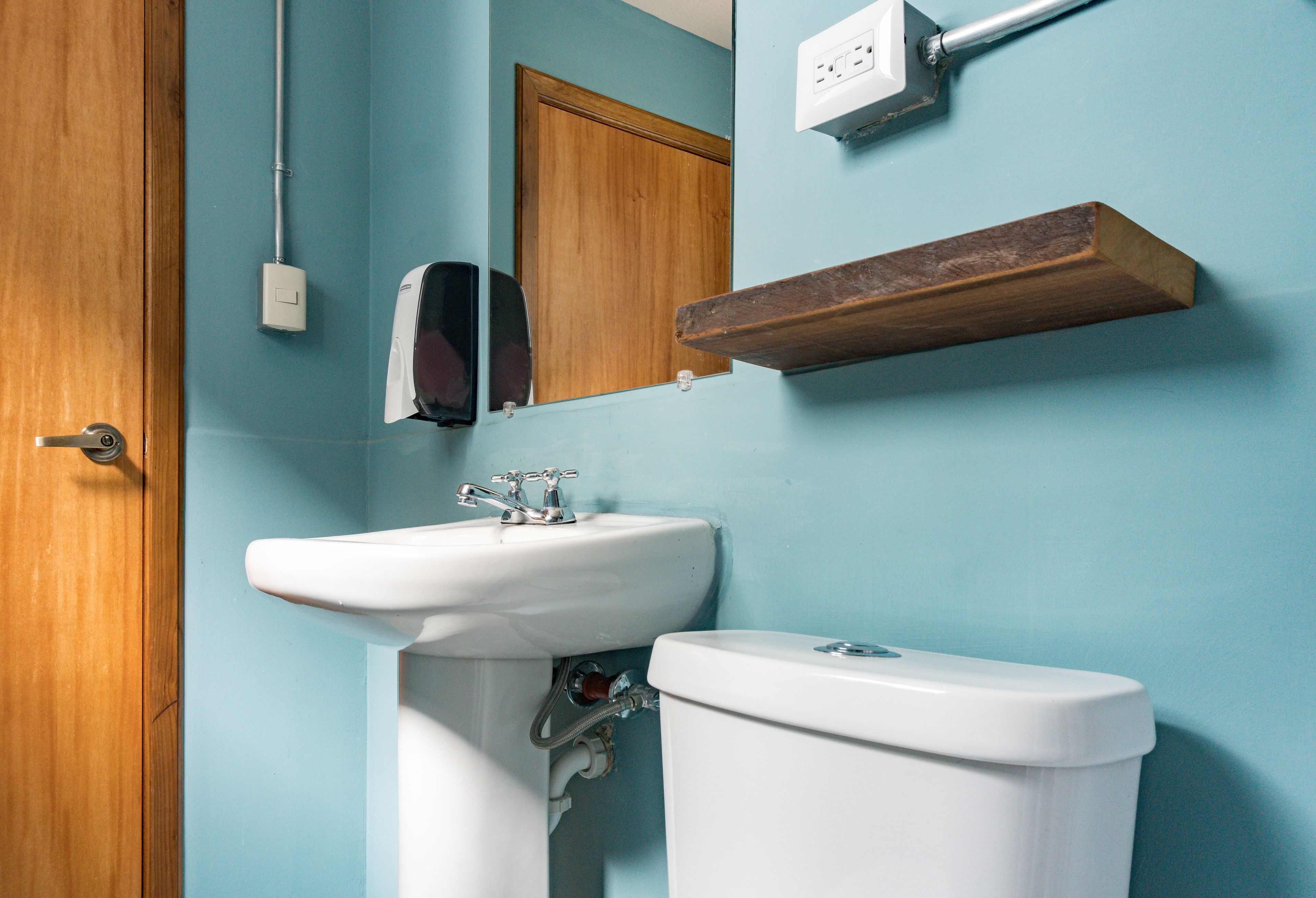 Une salle de bain avec une porte en bois, un lavabo blanc, un miroir et une étagère en bois au-dessus du lavabo.