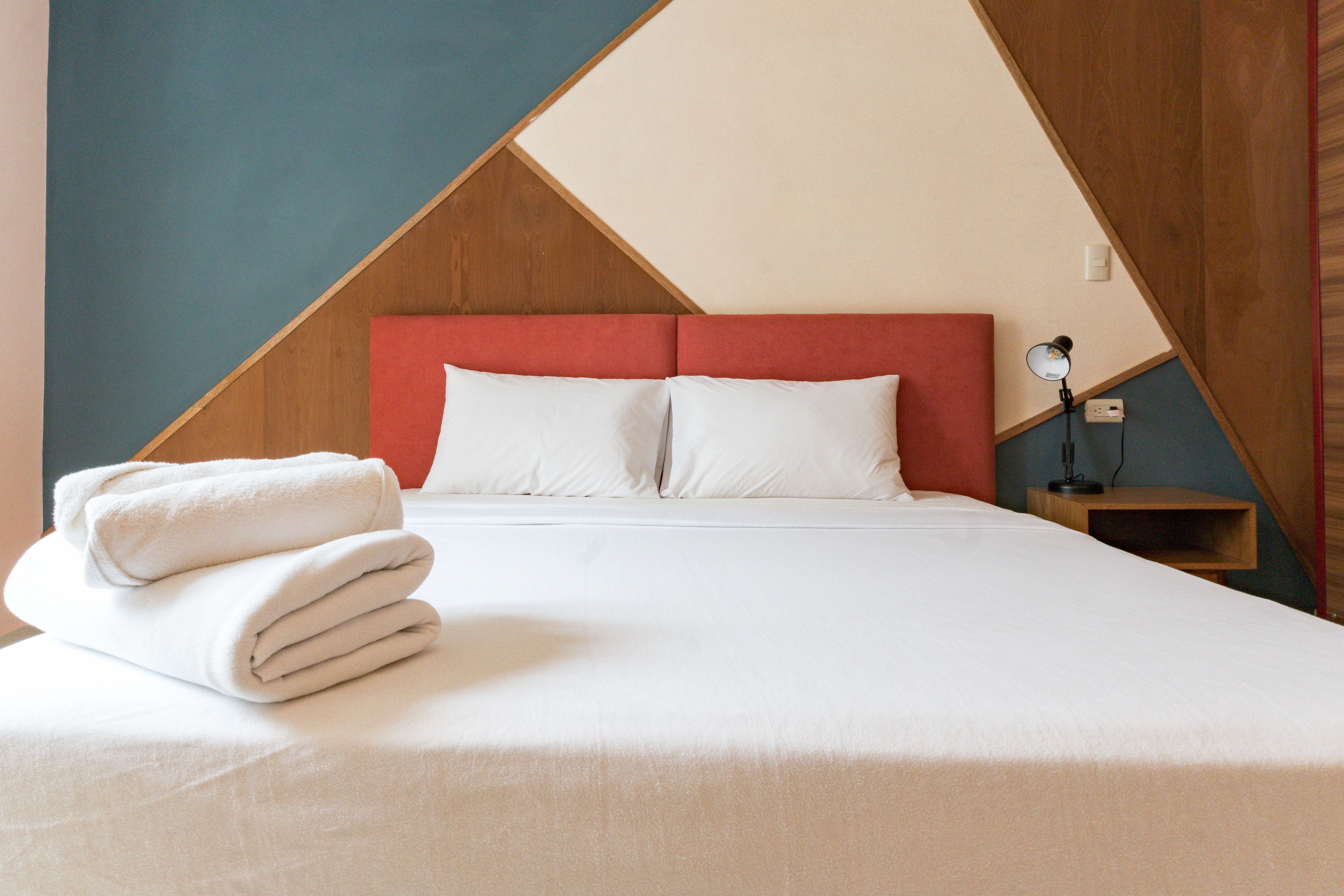 A neatly made bed with white linens and a folded towel, a wooden headboard, and a bedside table with a lamp.