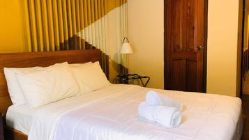A neatly made bed with white linens and a rolled towel on top, a wooden headboard, a lamp on a side table, and a wooden door.