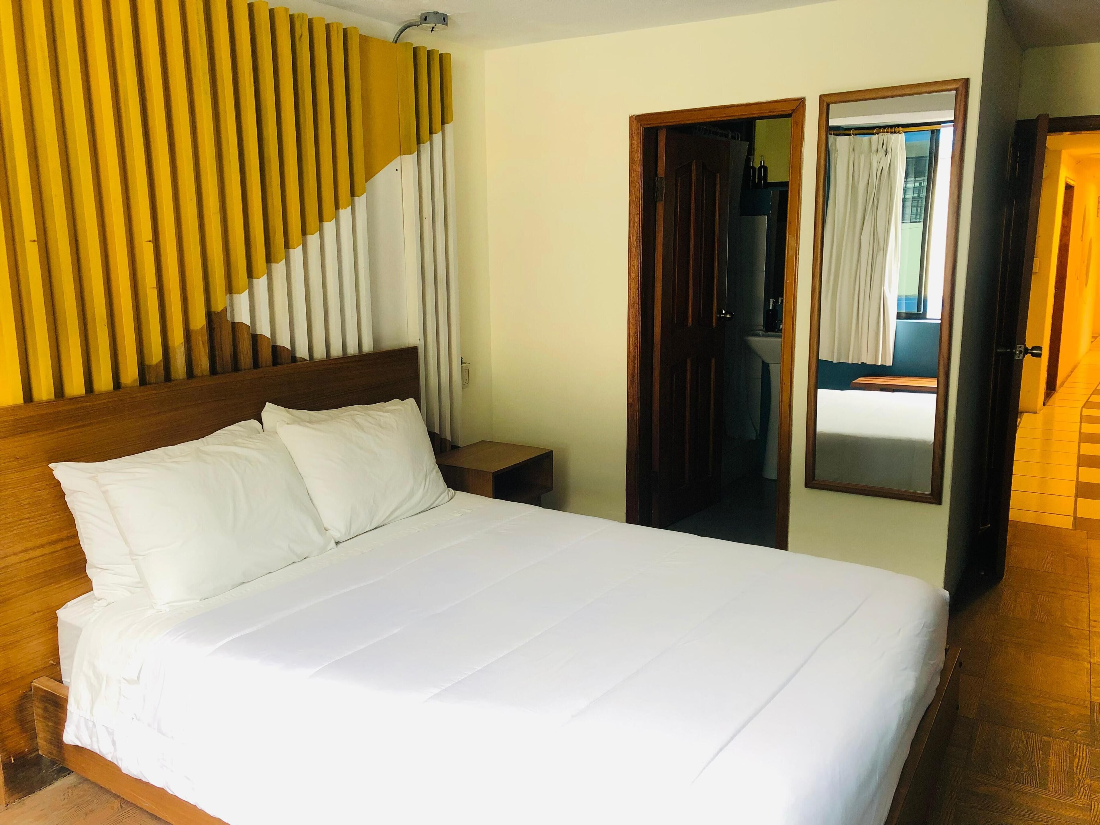 A hotel room with a large bed, wooden headboard, a nightstand, a mirror, a wooden staircase, and a bathroom visible through an open door.