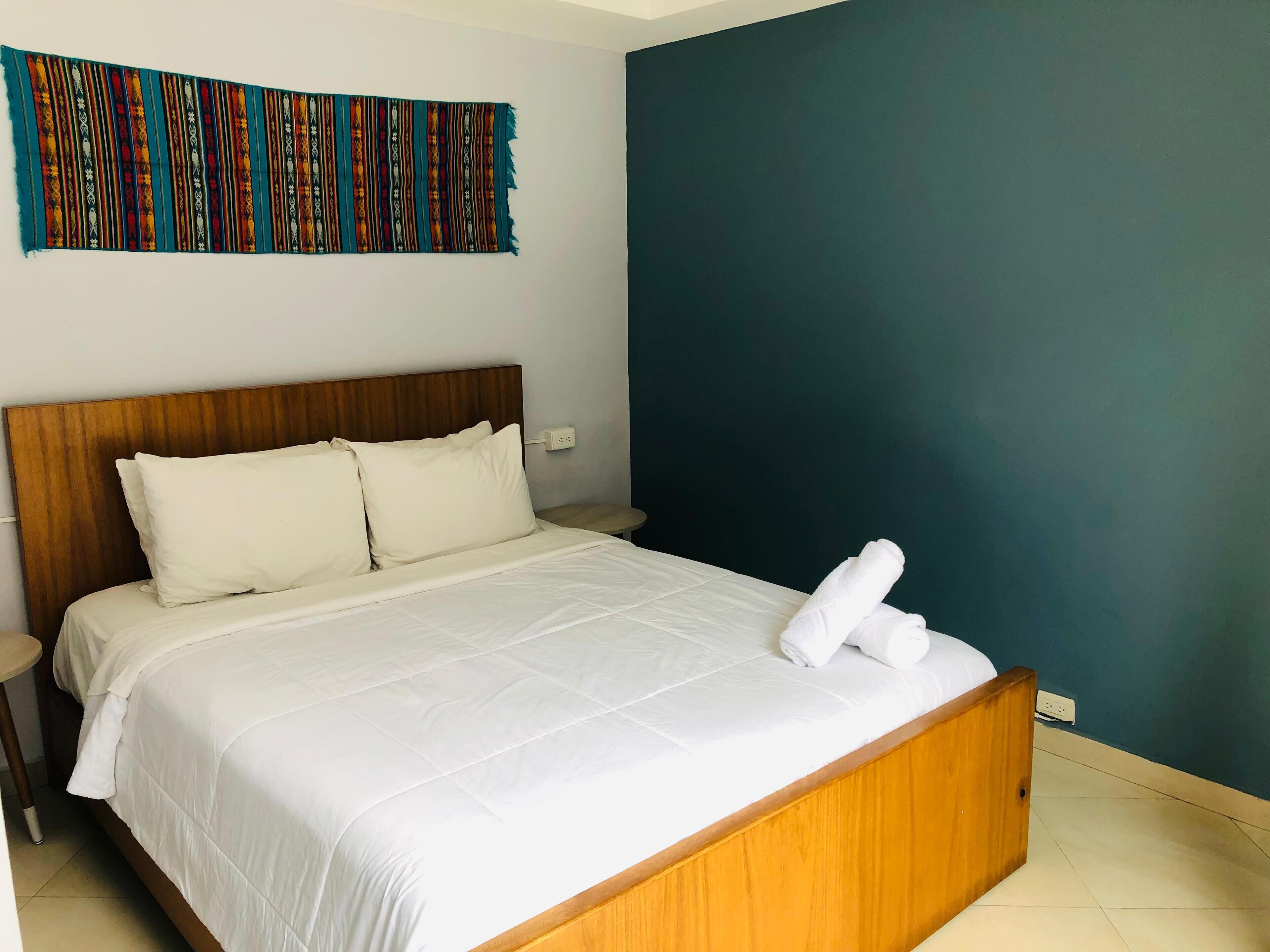 A neatly made bed with white linens and pillows, a wooden headboard, and a colorful tapestry on the wall.