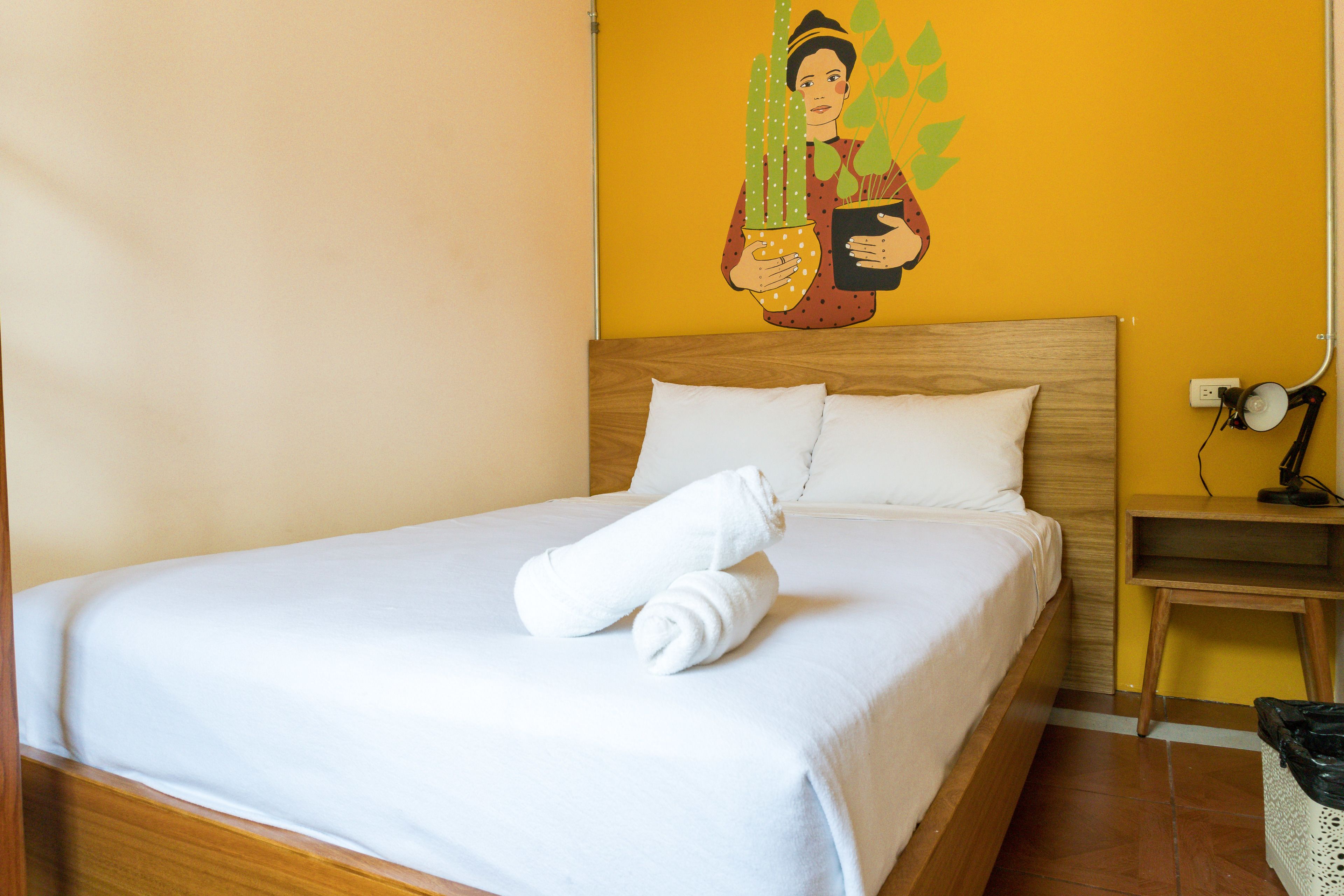 A neatly made bed with white linens and two rolled towels, a wooden headboard, a wall mural, a bedside table, and a lamp.