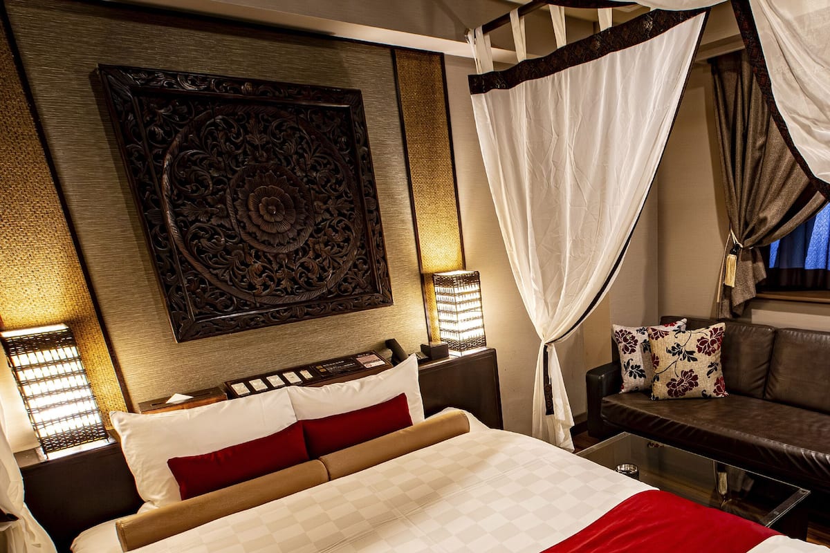 A hotel room with a bed, a sofa, a chair, a decorative wall panel, and a window with curtains.