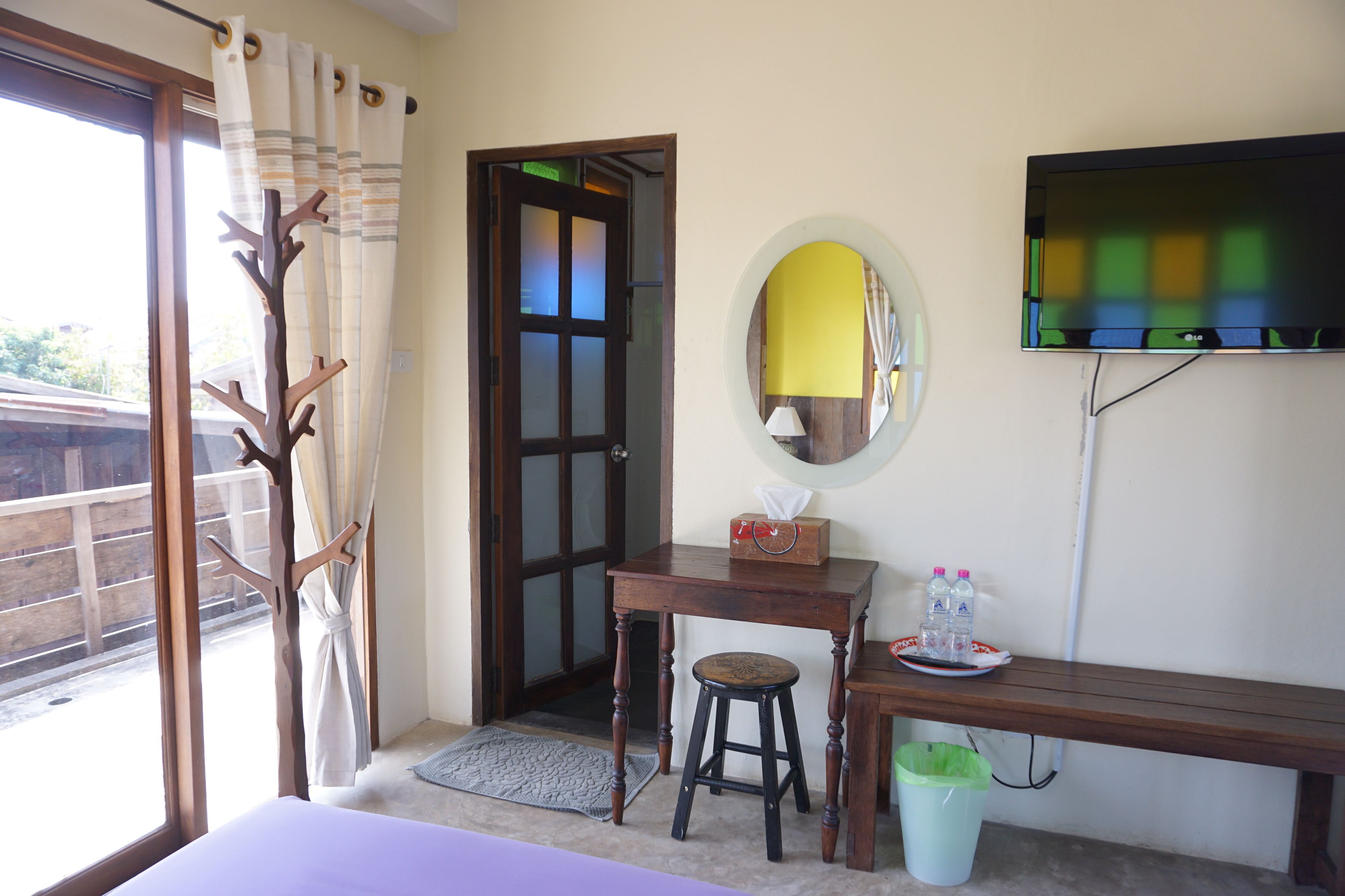 A room with a bed, a wooden desk with a stool, a trash bin, a television mounted on the wall, and a door leading outside.