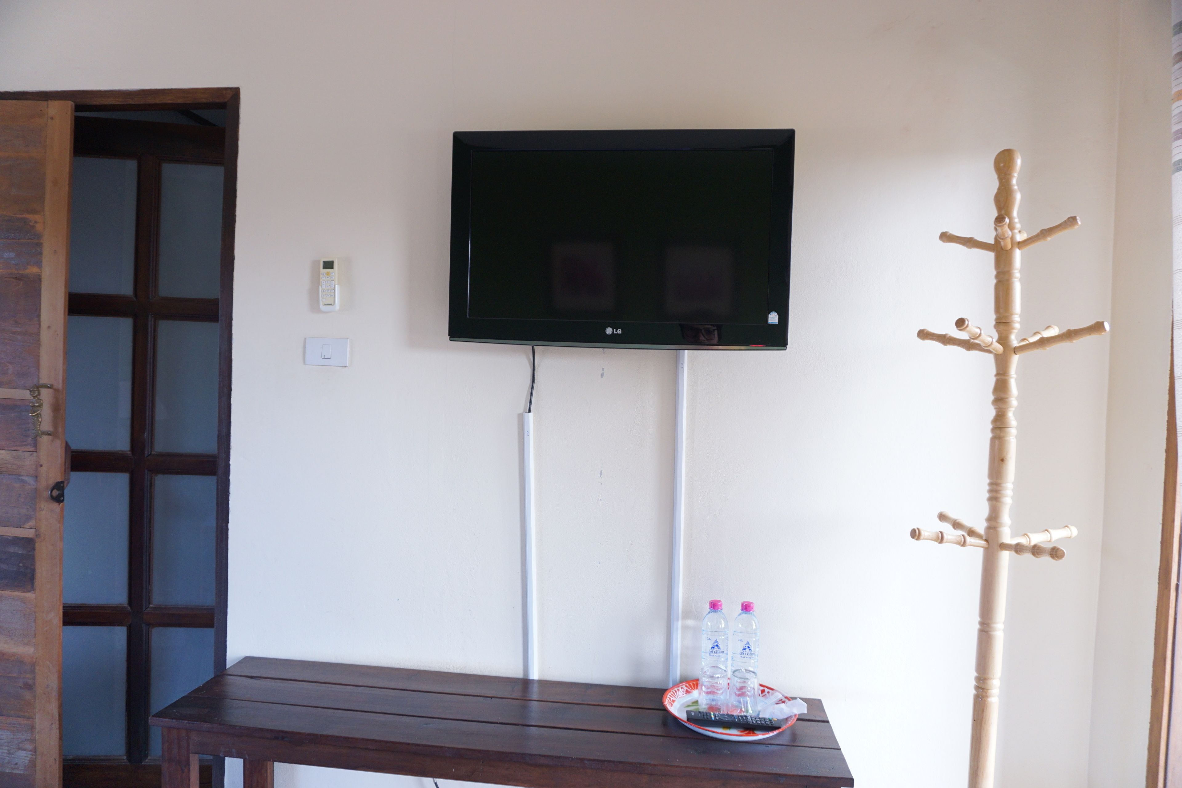 Una stanza con una TV a schermo piatto montata a parete, un appendiabiti in legno e un tavolino con telecomando e bottiglie d'acqua.