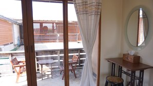 A room with a sliding glass door leading to an outdoor patio area. There is a wooden table and chairs outside, and a small stool inside the room. A mirror hangs on the wall, and a tissue box is on a small table. - Chiangkhanburi (Chiang Khan)