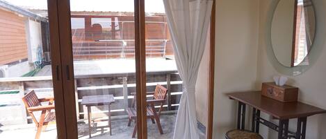 Habitación con puerta de vidrio corrediza que da a un patio exterior. Hay una mesa y sillas de madera afuera, y un taburete pequeño dentro de la habitación. Un espejo está colgado en la pared, y una caja de pañuelos sobre una mesita.
