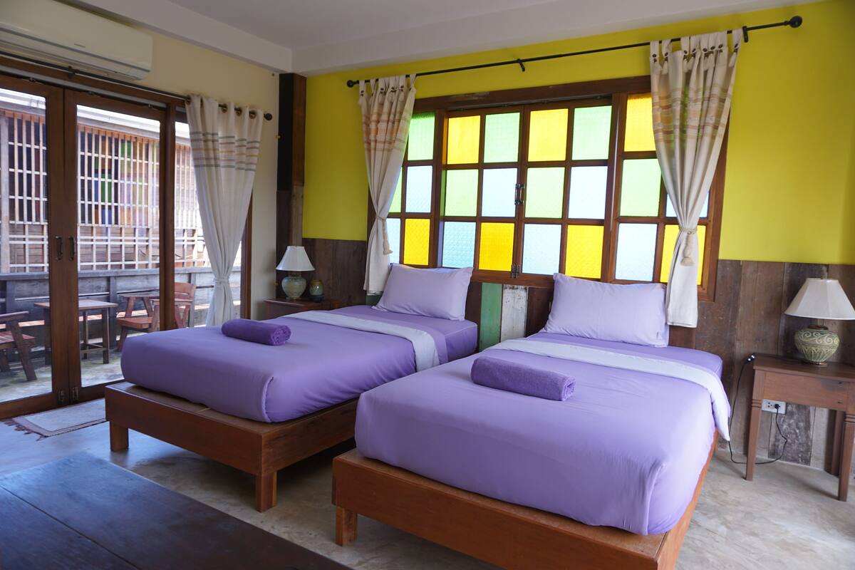 A room with two beds, a wooden floor, a window with curtains, and a view of the outside.
