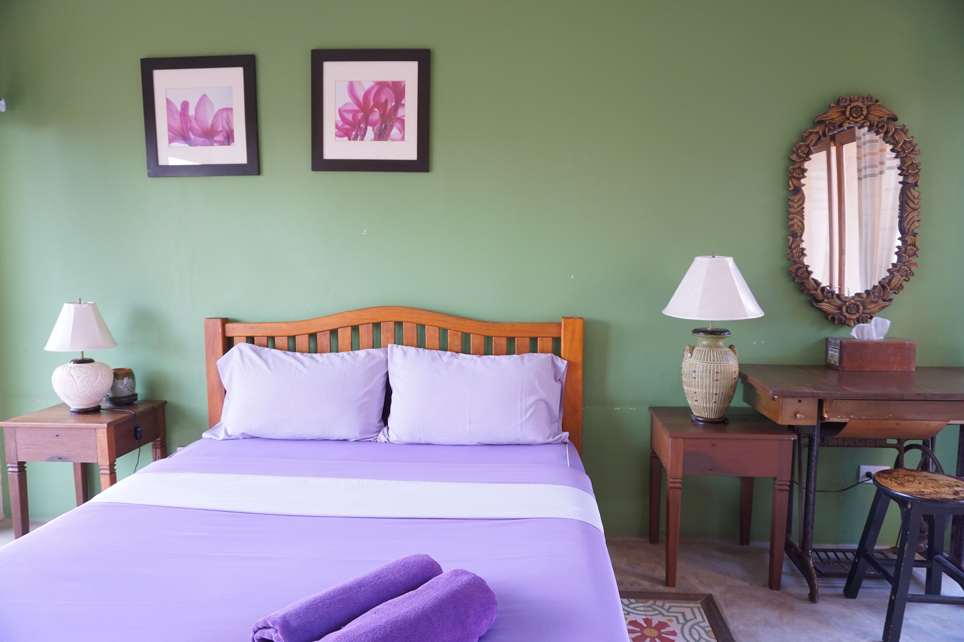 A neatly made bed with a wooden headboard, two bedside tables with lamps, a desk with a mirror, and two framed floral pictures on the wall.