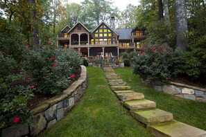 Une grande maison à étages, avec un chemin de pierre menant à l'entrée, entourée d'une végétation luxuriante et de plantes en fleurs.