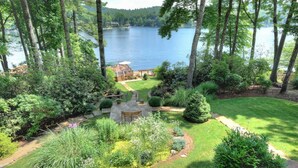 A lush garden with a patio area, surrounded by tall trees and a lake view.