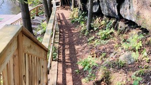 A wooden deck with a metal railing overlooking a forested area with a rocky cliff and a path leading into the woods.