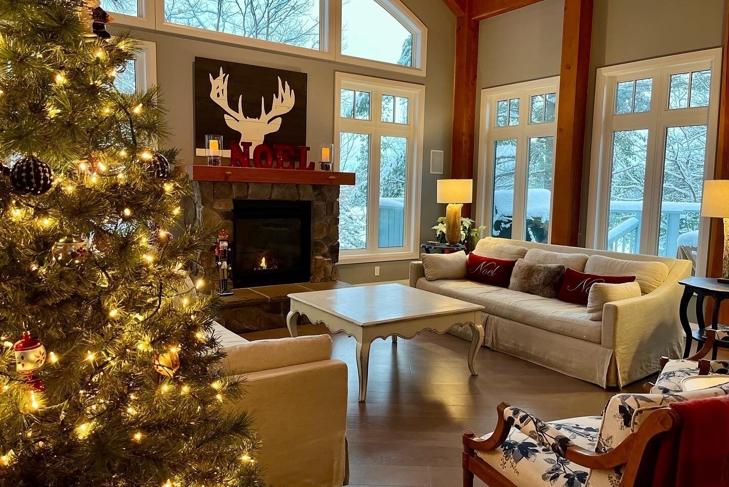 A cozy living room with a fireplace, a Christmas tree, and a large window with a view of snow-covered trees.