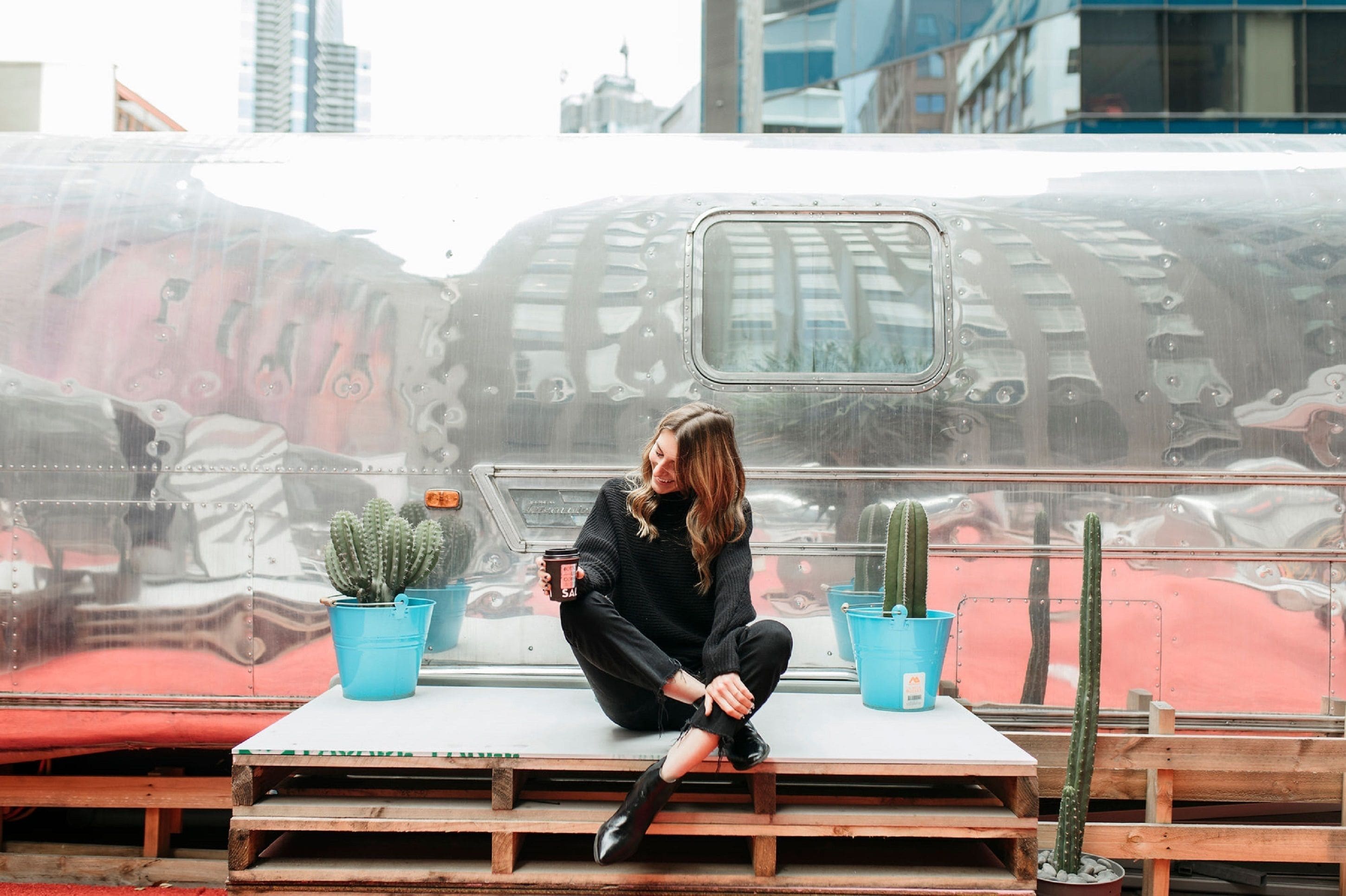 A person sitting on a bench with potted cacti and a beverage, in front of a large, reflective vehicle.
