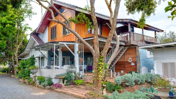 A two-story house with a wooden exterior, a balcony, and a garden with various plants and a small statue. - Rosemary House (Pak Chong)