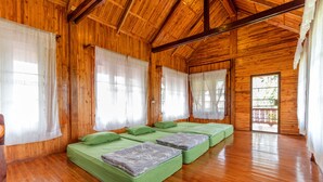 A wooden cabin with four green beds arranged in a row, large windows with sheer curtains, and a wooden floor. - Rosemary House (Pak Chong)