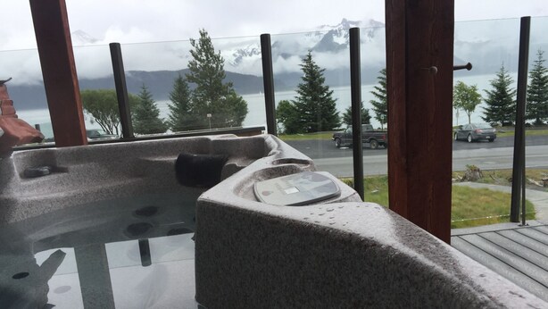 Una bañera de piedra con cerramiento de vidrio, con vistas a una carretera y un cuerpo de agua con montañas al fondo.
