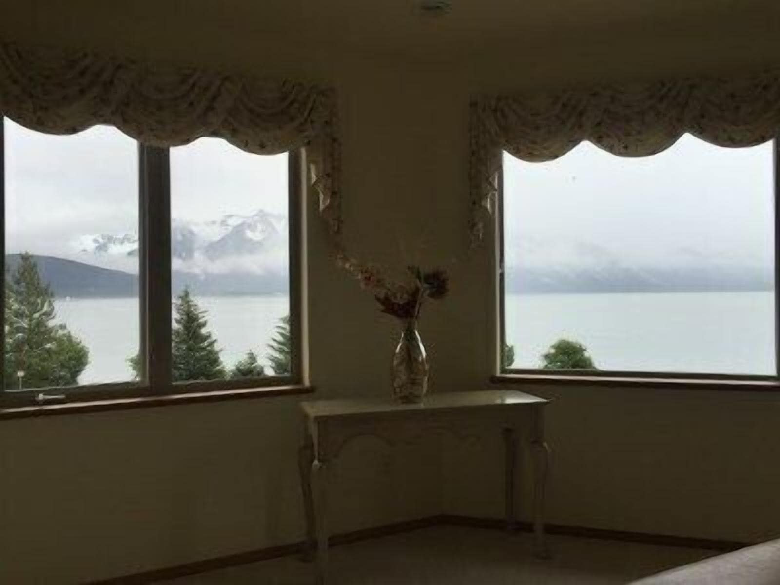 Une pièce dotée de deux fenêtres donnant sur un lac et des montagnes, une petite table sur laquelle est posé un vase de fleurs, et un mur sobre.