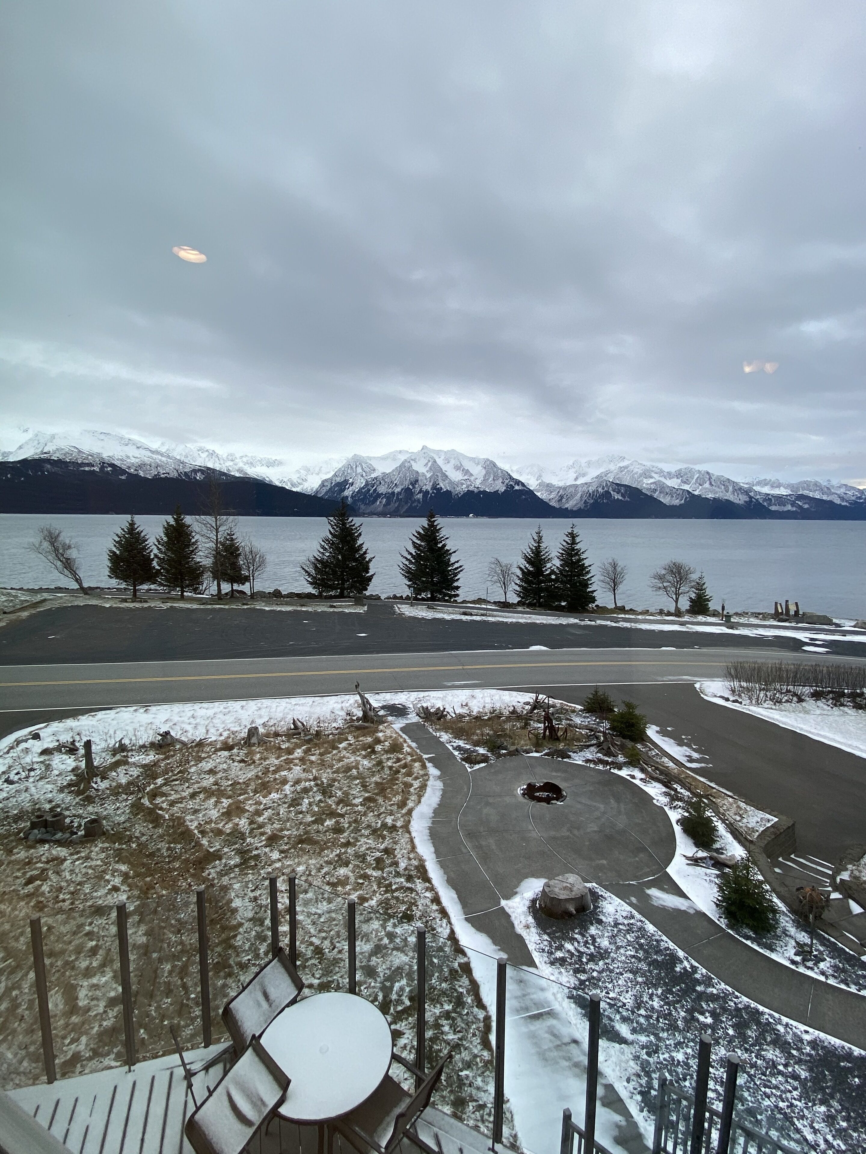 A lakeside view with snow-covered mountains, a paved area with a fire pit, and a wooden deck with chairs.