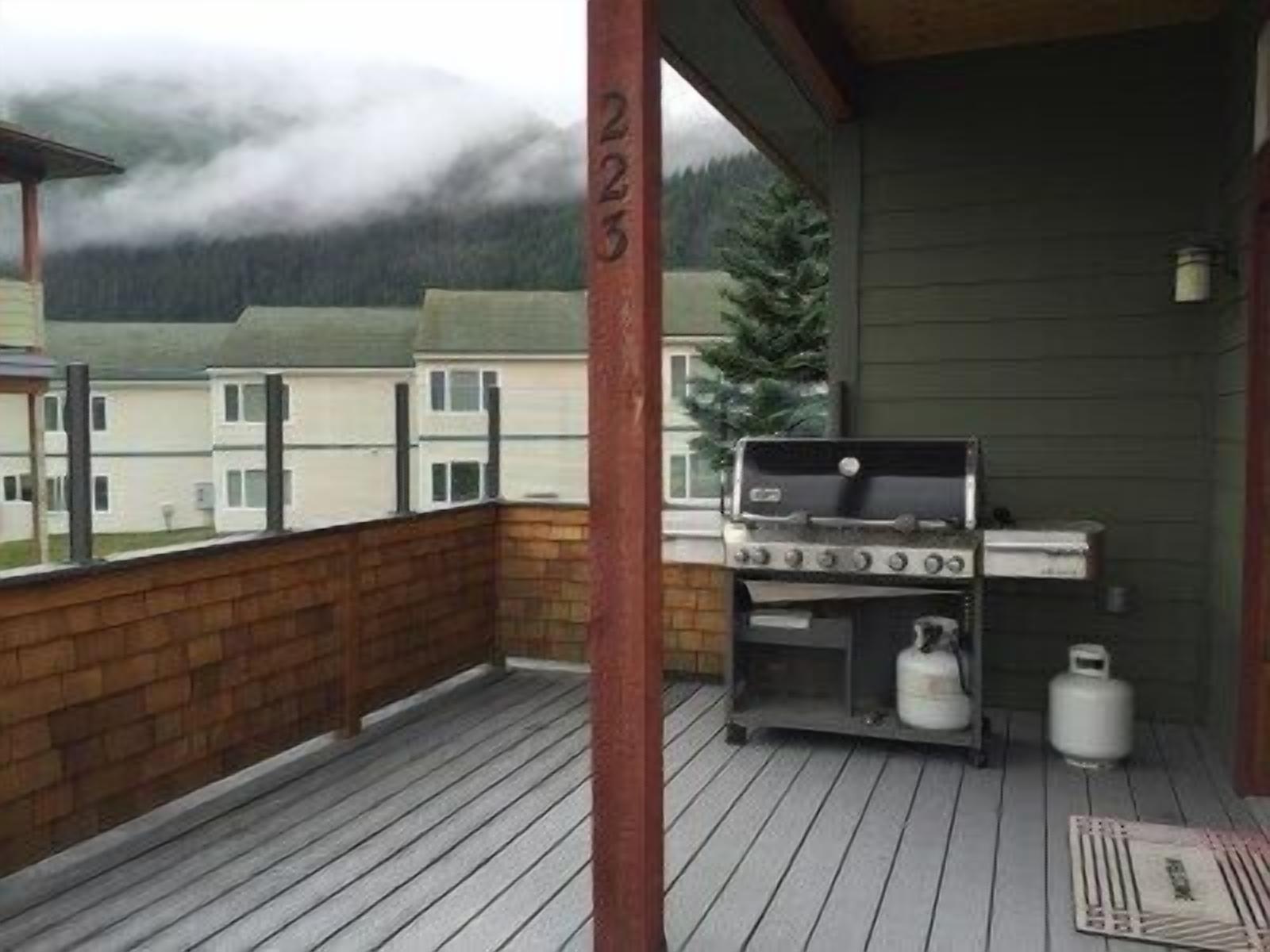 A covered balcony with a barbecue grill, a propane tank, and a view of neighboring buildings and mountains.