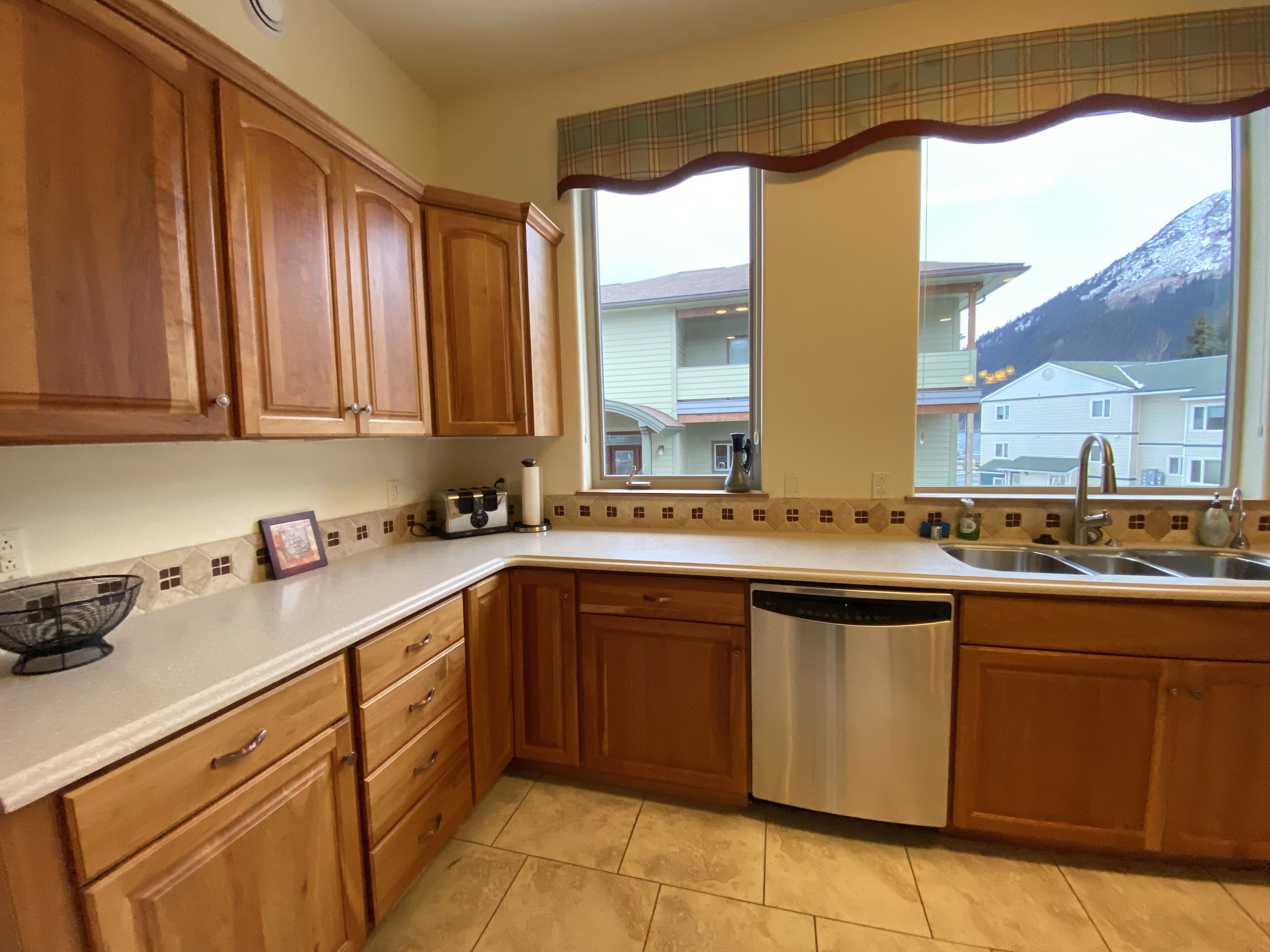 A kitchen with wooden cabinets, a dishwasher, a window with a view of houses and mountains, and a tiled backsplash.