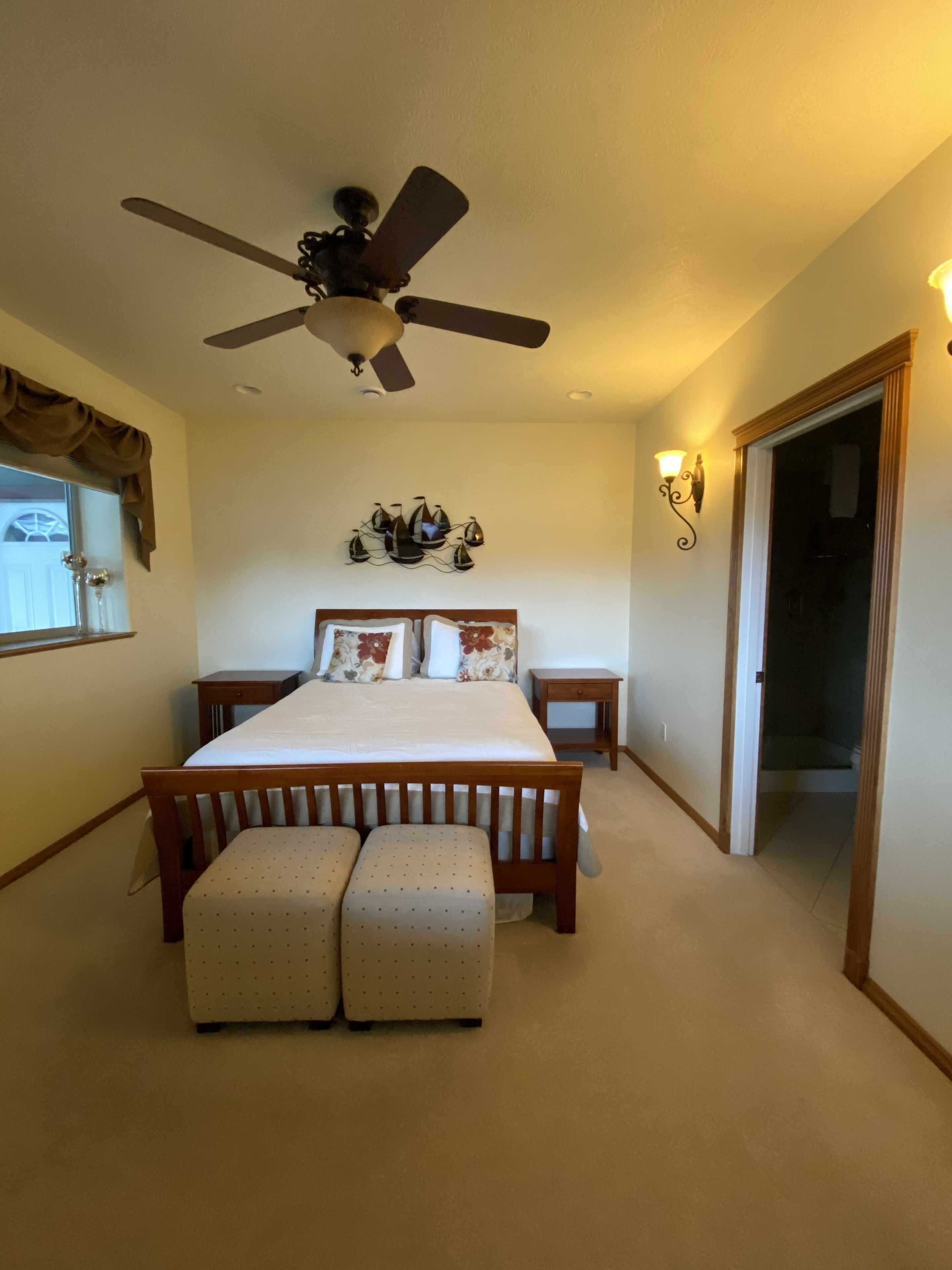 A bedroom with a bed, two nightstands, a ceiling fan, a wall-mounted light fixture, and a window with curtains.