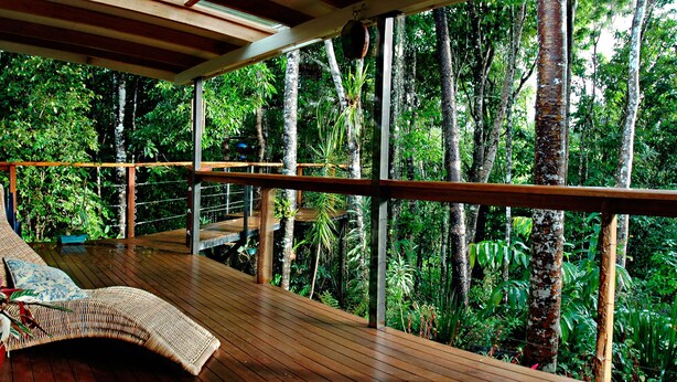 A wooden deck with a wicker lounge chair and a small table, surrounded by lush greenery and trees. - Sharlynn by the River (Malanda)
