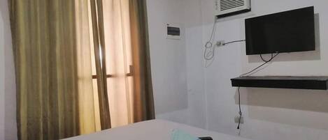 A hotel room with a bed, a TV mounted on the wall, an air conditioning unit, and a window with curtains.