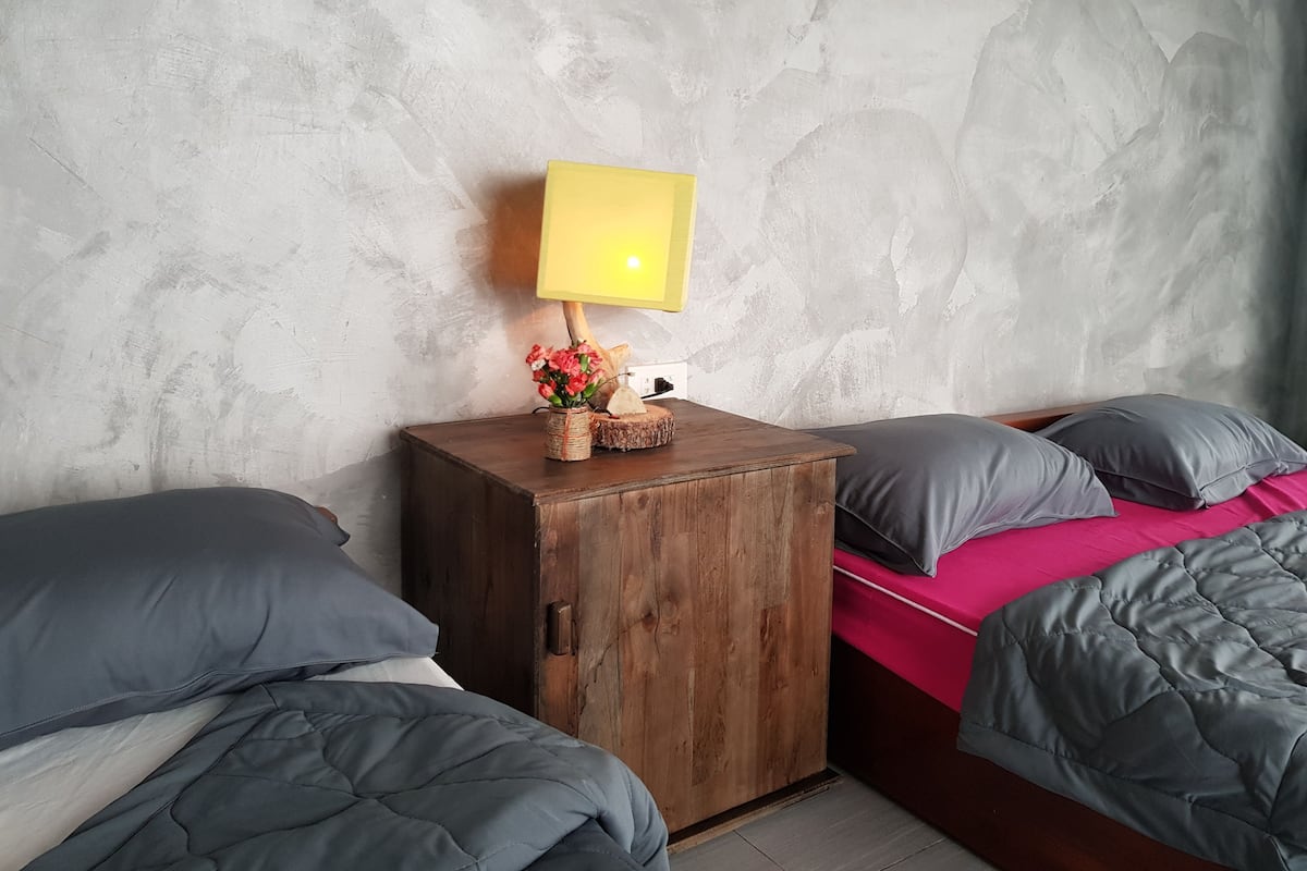 Un dormitorio con un armario de madera, una cama con ropa de cama gris, una manta rosa y una mesita de noche con una lámpara y flores.
