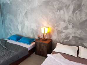A room with two beds, one with blue and white bedding and the other with white bedding. There is a nightstand with a lamp and a small vase with flowers. - Confetti House (Quy Nhon)