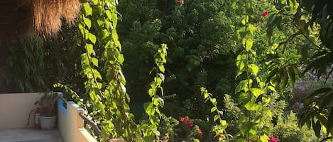 A balcony with a thatched roof, potted plants, and a view of lush greenery and pink flowers.