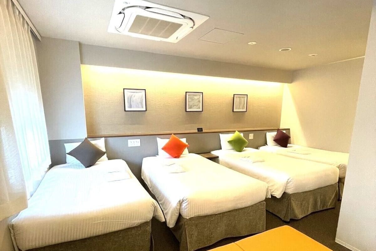 A hotel room with two beds, a bench, a ceiling light, and framed artwork on the wall.